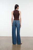 A model shown from the back wearing a dark brown sleeveless top and wide-leg blue jeans, standing in a minimalist studio with a white wall and concrete floor.