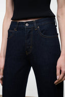 Close-up of a model wearing dark indigo denim jeans with a black ribbed top, showing the front waistband, button, and pocket details.