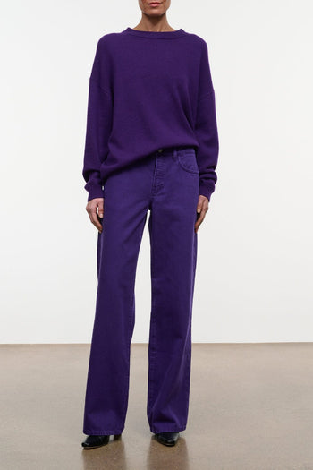 A model wearing a matching purple lounge set consisting of a crewneck sweatshirt and wide-leg trousers, standing against a plain white wall.