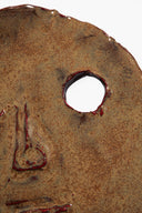 Ceramic face ornament detail: circular hole and painted nose/mouth on dark brown glaze.